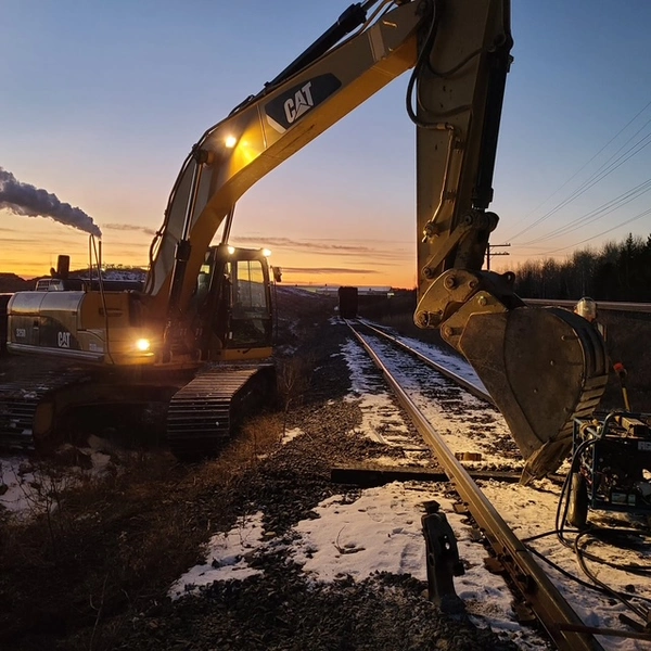 A CAT excavator works beside railway tracks at sunset, with smoke rising in the background and snow covering the ground.